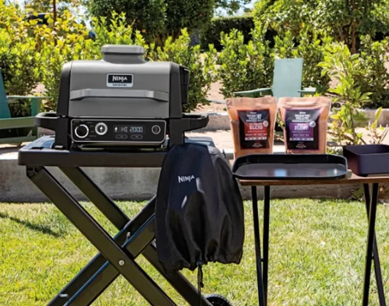 Ninja Woodfire grill on its stand outdoors with the weather-proof cover and pellet blends